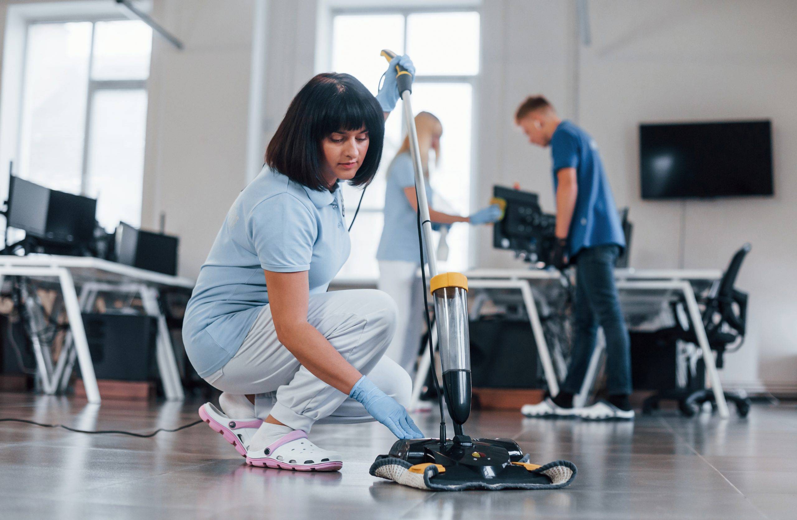 Woman uses vacuum cleaner. Group of workers clean modern office together at daytime.